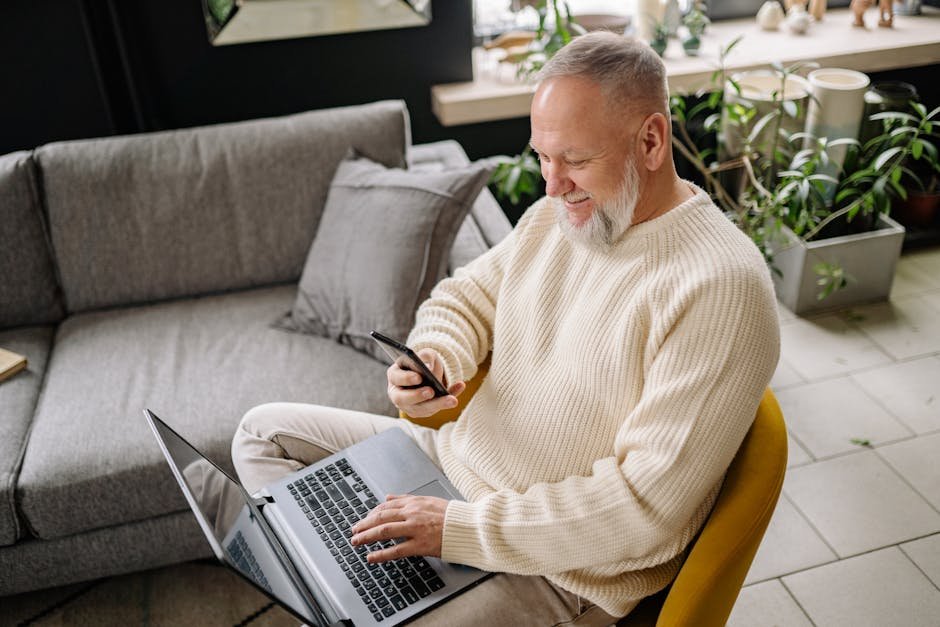 A smiling elderly man sits comfortably indoors, using both a phone and laptop, with a cozy ambiance.