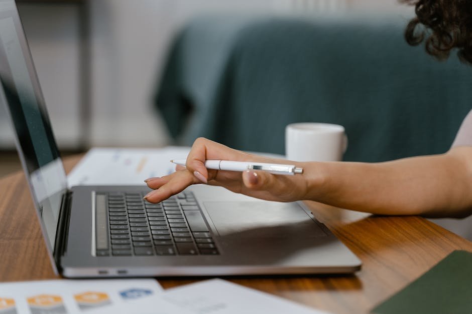 A woman works remotely on her laptop while holding a pen, focused on her task in a casual home setting.