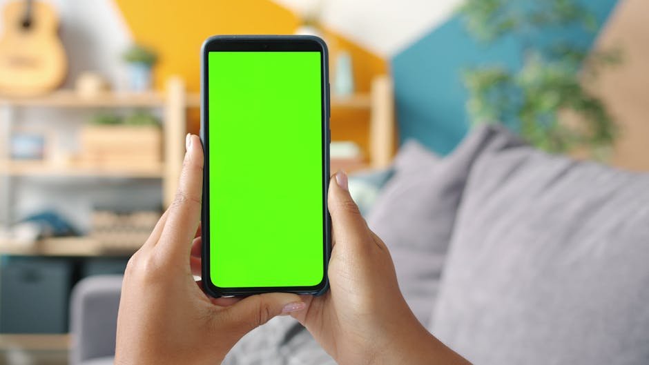 Hands holding smartphone with green screen in modern living room.