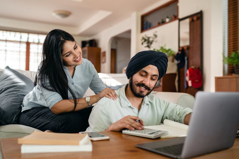 Positive ethnic man sitting at coffee table with laptop and smartphone and books and notebook and calculating while smiling ethnic wife sitting on sofa and supporting and watching behind